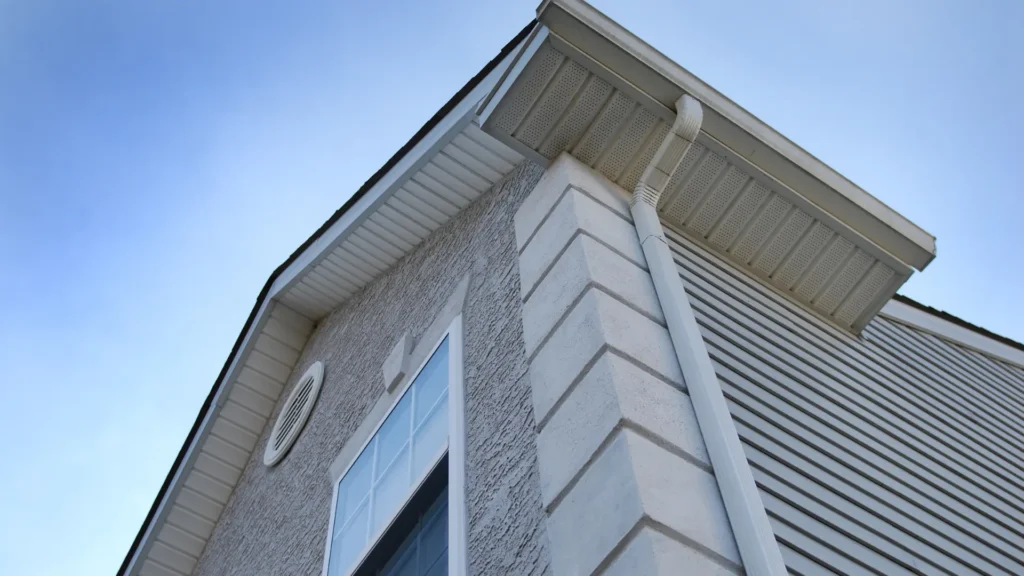 Textured stucco wall with a window and round vent, under vented soffit and a white gutter and downspout